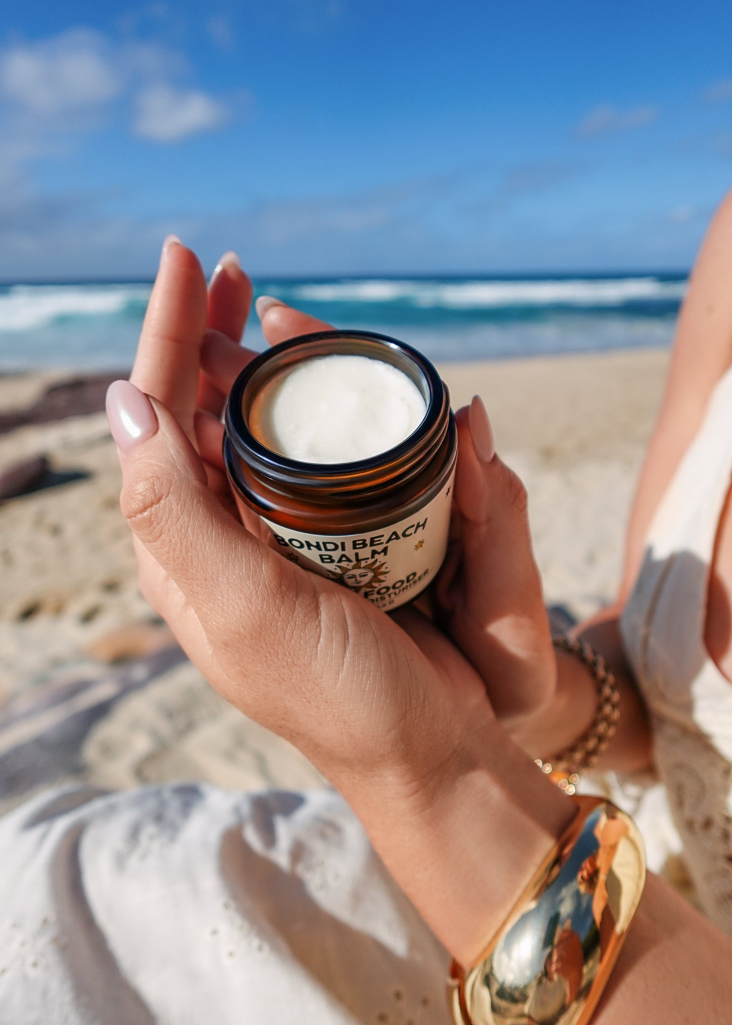 Person holding a jar of 'Bondi Beach Balm' Face Food on a beach with ocean view