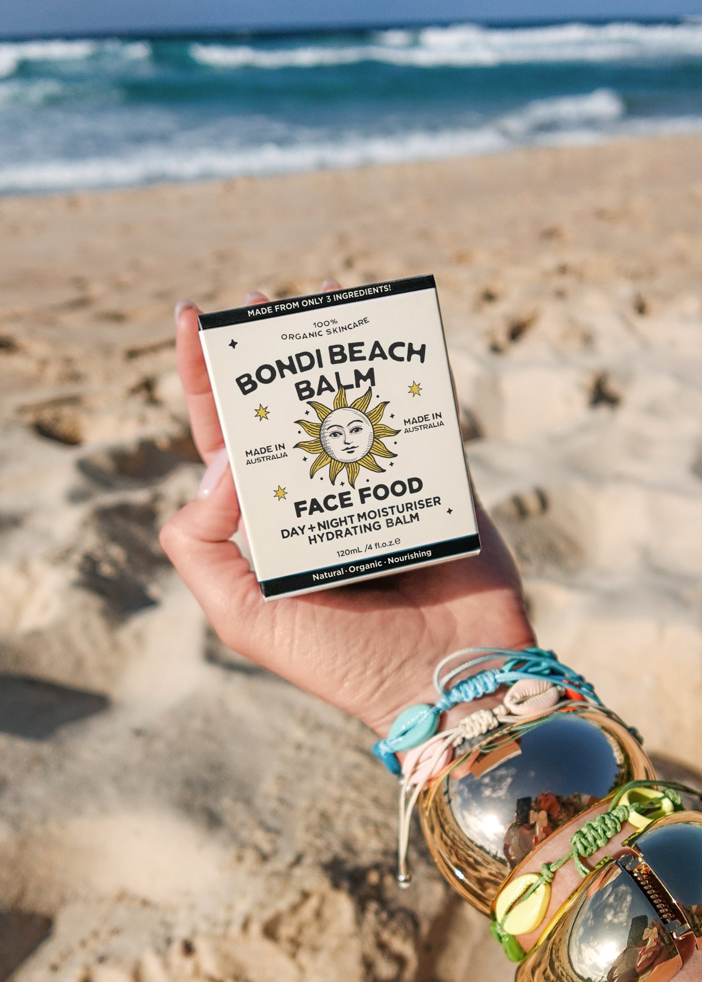 Hand holding a 'Bondi Beach Balm' Face Food product on a sandy beach with ocean waves in the background