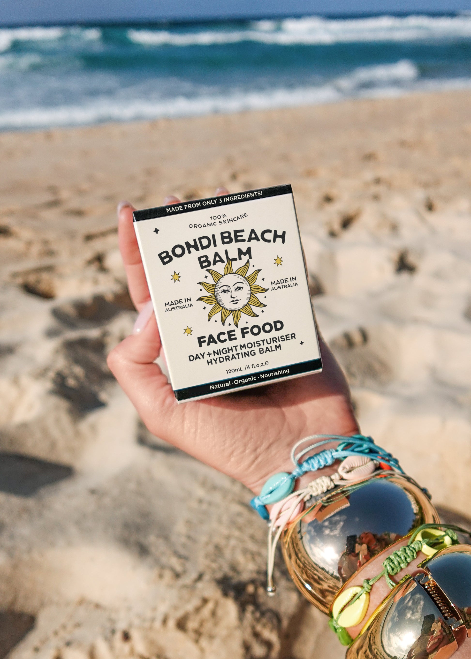 Hand holding a 'Bondi Beach Balm' Face Food product on a sandy beach with ocean waves in the background