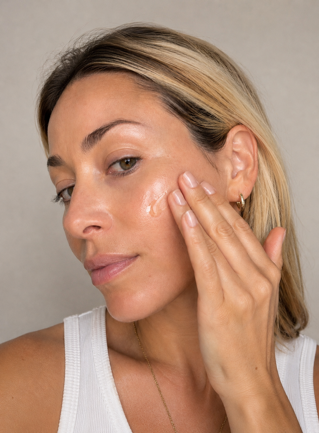 A close-up of a woman with blonde-highlighted hair applying a castor oil to her cheek, wearing a white ribbed tank top against a neutral beige background.